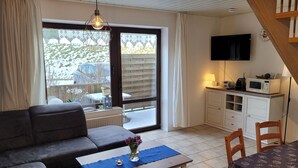 Una sala de estar con un sofá, una mesa de centro con un florero, una mesa de comedor con sillas, un televisor y una zona de cocina con armarios y un microondas.