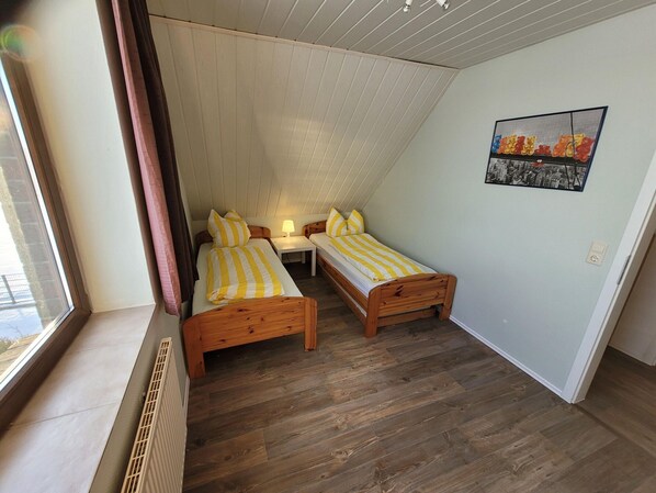 A small, cozy room with two wooden beds, a window, a radiator, and a cityscape painting on the wall. - Apartment in Nebmersiel mit Terrasse (Dornum)