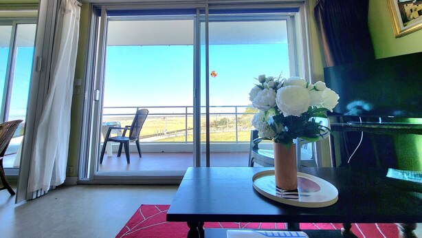 Una habitación con balcón, una mesa con un florero, una silla y un televisor.