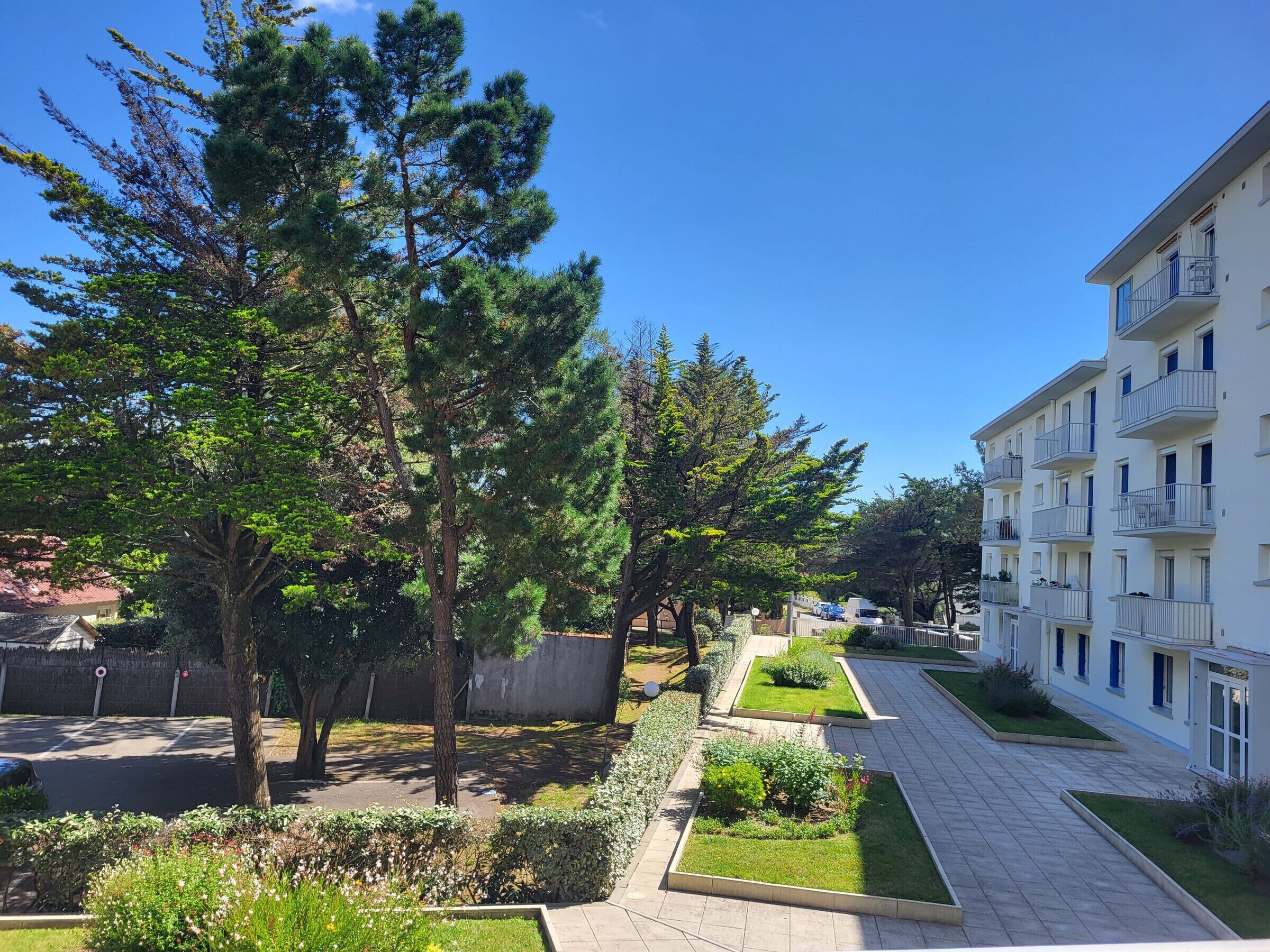 A residential area with a paved walkway, trees, and a multi-story building with balconies.