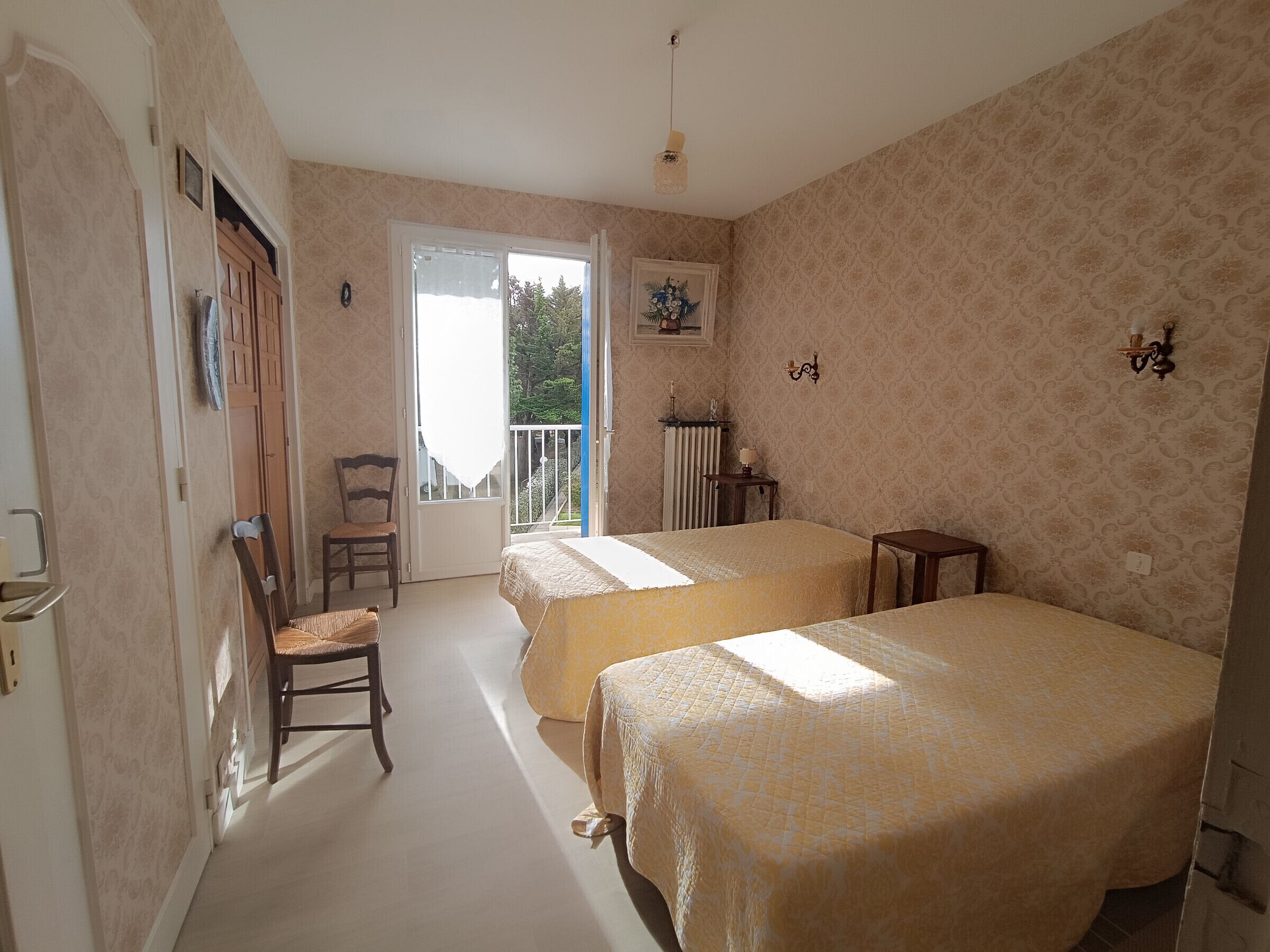 A room with two beds, a chair, a radiator, and a balcony with a view of trees.