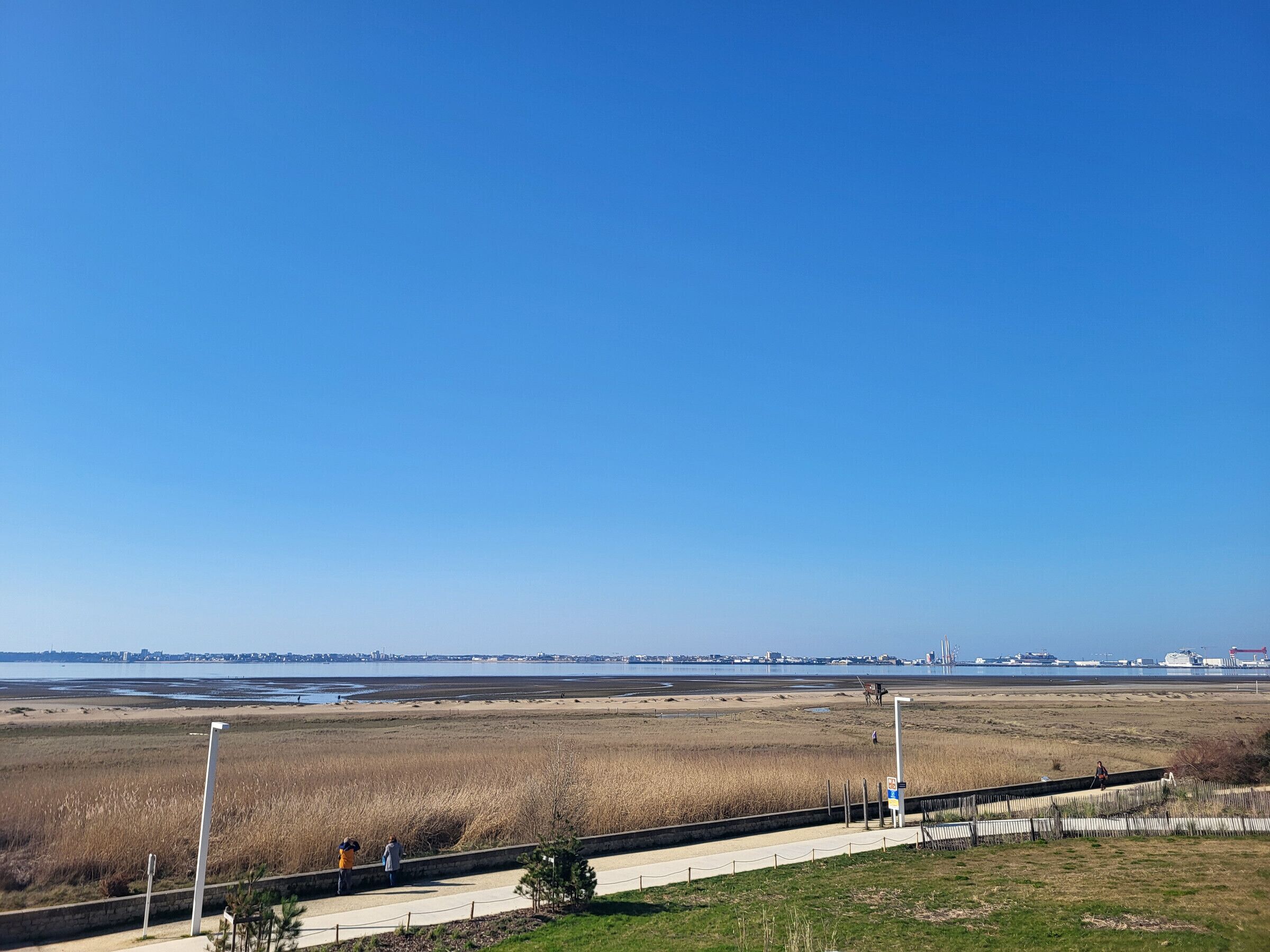 A coastal landscape with a clear sky, a sandy beach, and a distant city skyline.