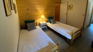 A small, cozy room with two single beds, a nightstand with a lamp, wooden paneling on the walls, and framed pictures on the wall.