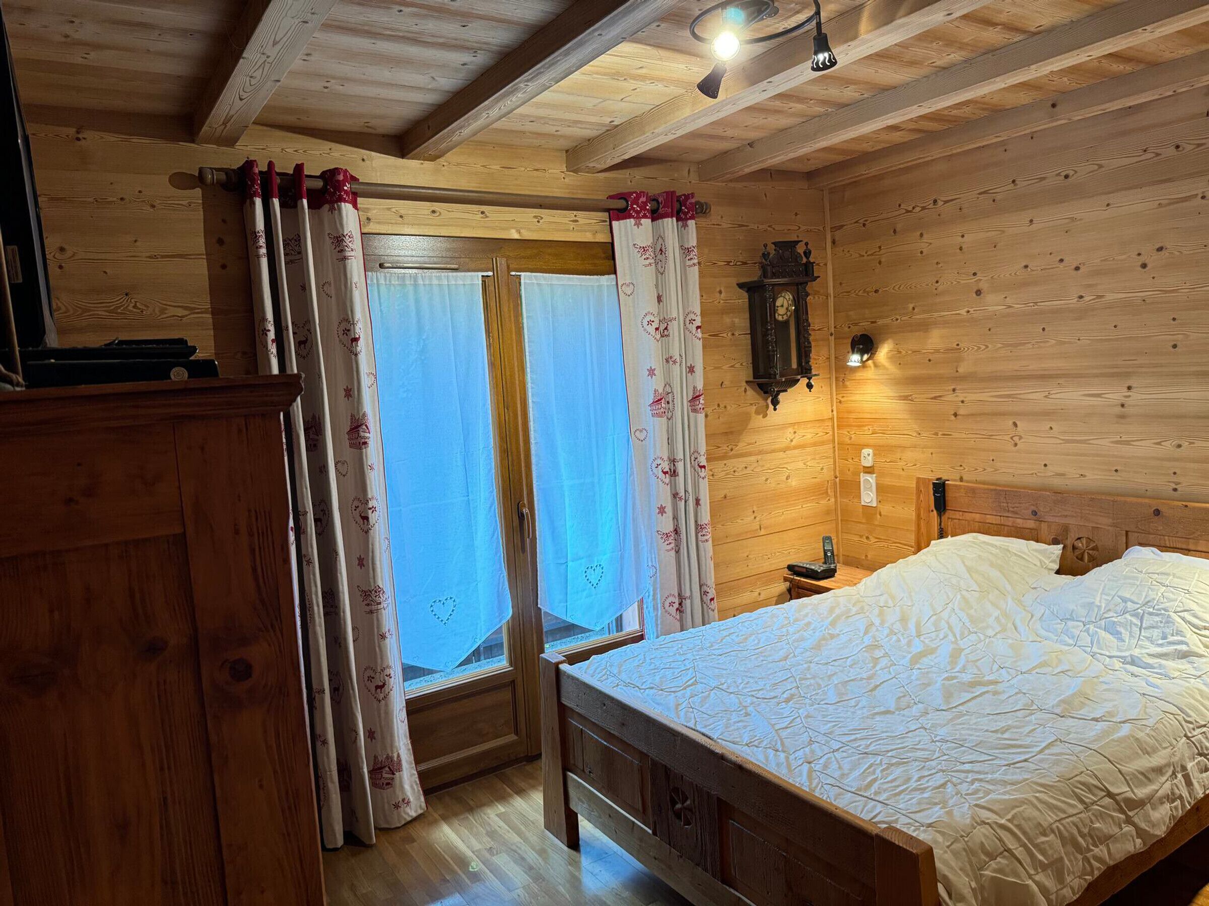 A wooden cabin bedroom with a bed, curtains, a clock, and a small table.