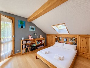 Quarto com teto inclinado, claraboia, cama de madeira, estante e porta para uma varanda.