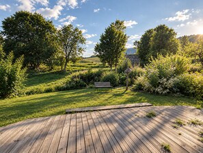 Um deck de madeira com um banco em um jardim exuberante, com árvores e arbustos.