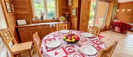 A dining area with a round table set for four, wooden chairs, and a kitchen area with wooden cabinets and a window overlooking a garden.