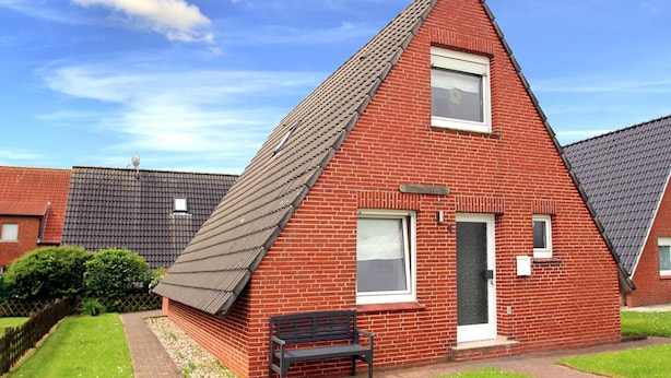 A red brick house with a sloped roof, a front door, and a bench in the front yard. - Ferienhaus Toms Hutte mit Terrasse in Dornum (Dornum)