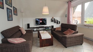 A living room with two brown sofas, a coffee table, a TV, and a window with a view of a house. - Ferienhaus Toms Hutte mit Terrasse in Dornum (Dornum)