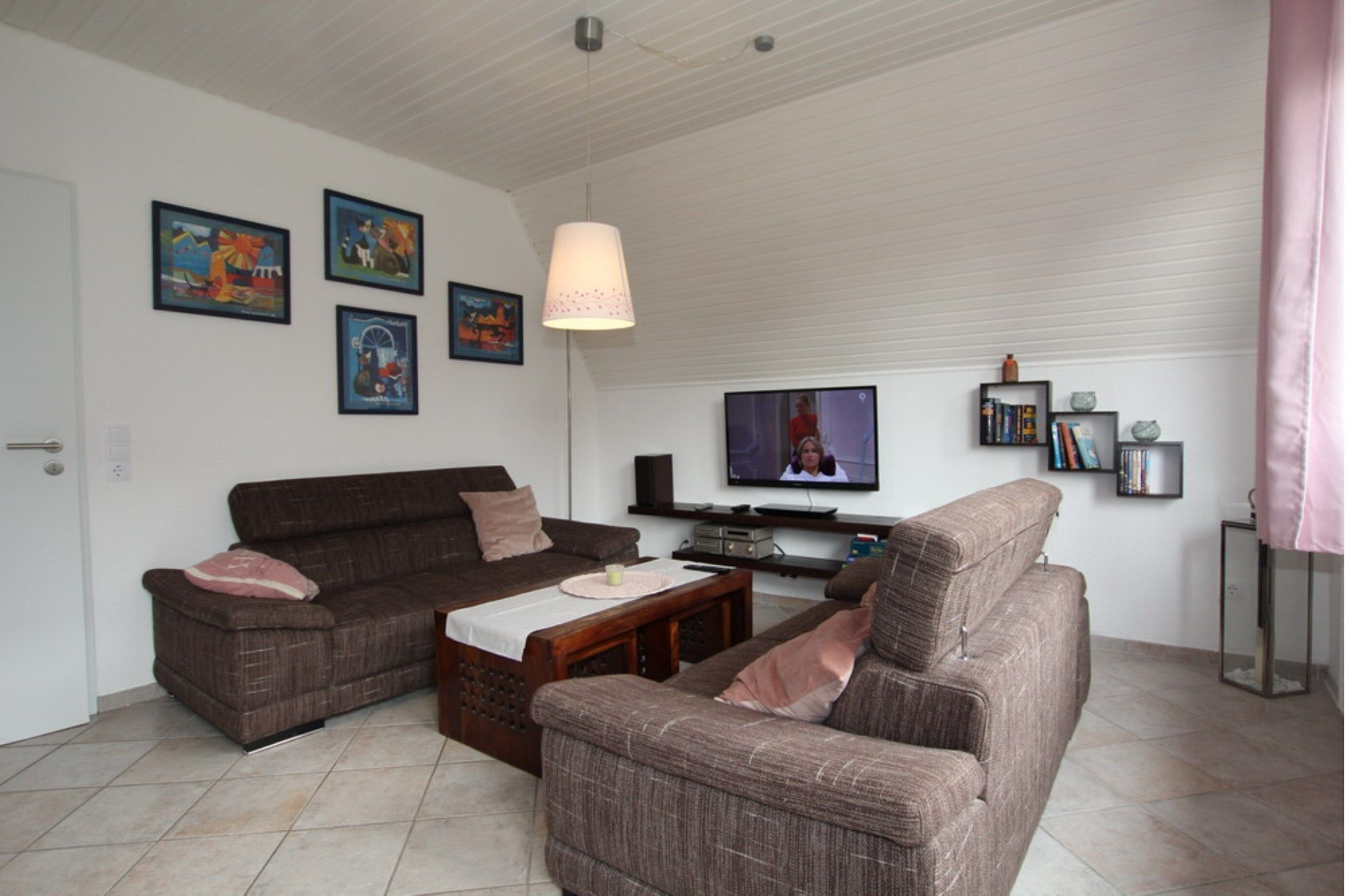 Una sala de estar con dos sofás marrones, una mesa de centro y un televisor. Hay cuadros enmarcados en la pared y una lámpara rosa.