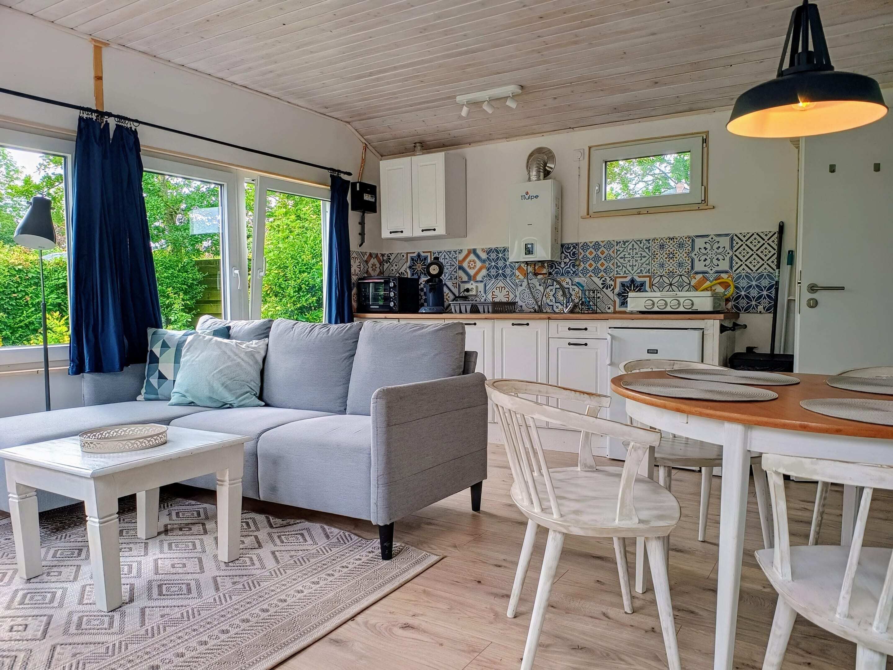 Una sala de estar acogedora con un sofá gris, una mesa de centro blanca y un comedor con sillas blancas y una mesa de madera. Se ve una cocina con armarios blancos y un salpicadero de azulejos al fondo.