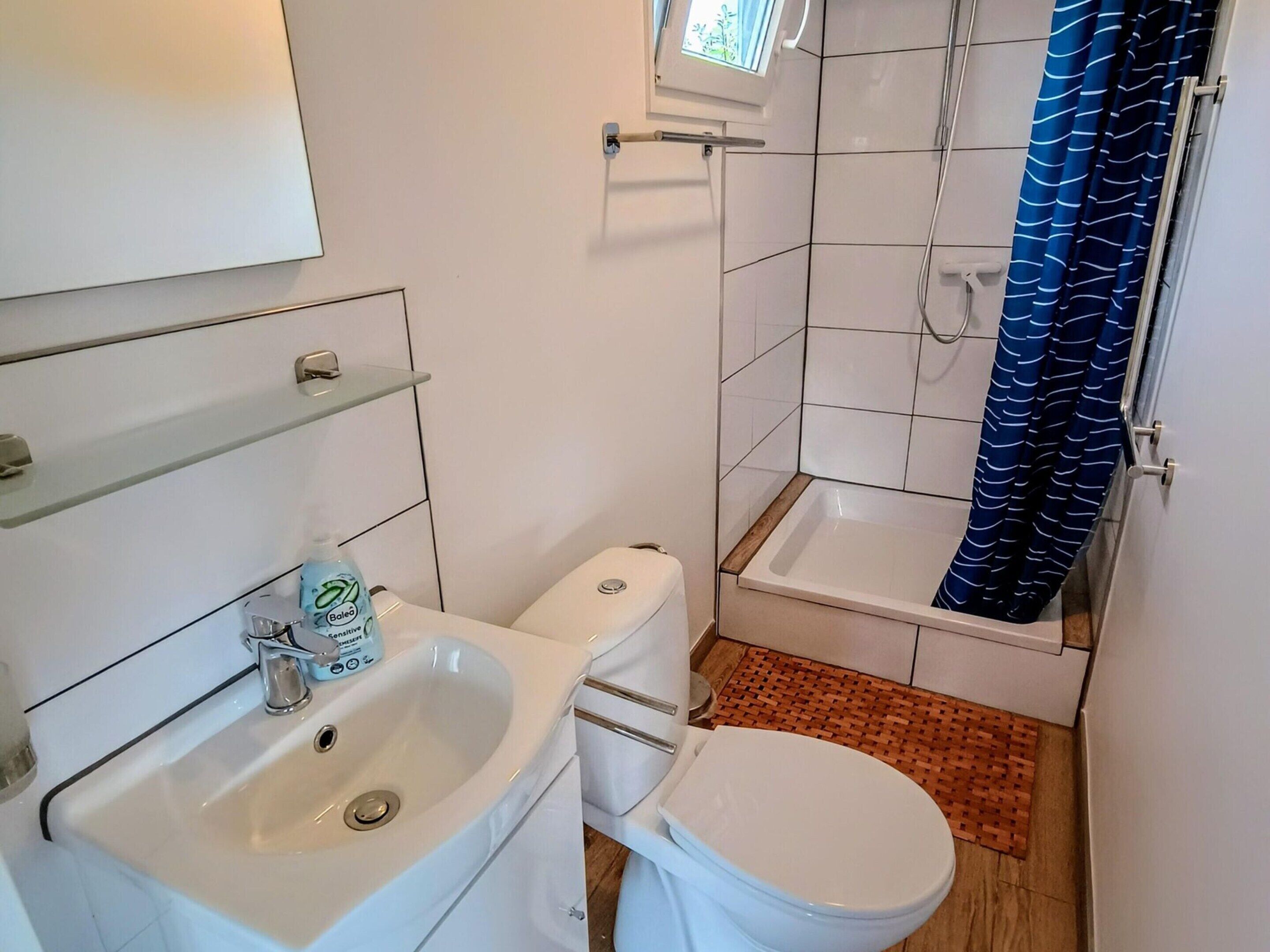 A bathroom with a white sink, toilet, and shower. There is a window above the shower, and a blue shower curtain.