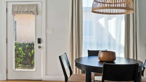 Un coin repas avec une table ronde, des chaises, un luminaire en fibres tressées et, vue par une porte, un balcon agrémenté de verdure.