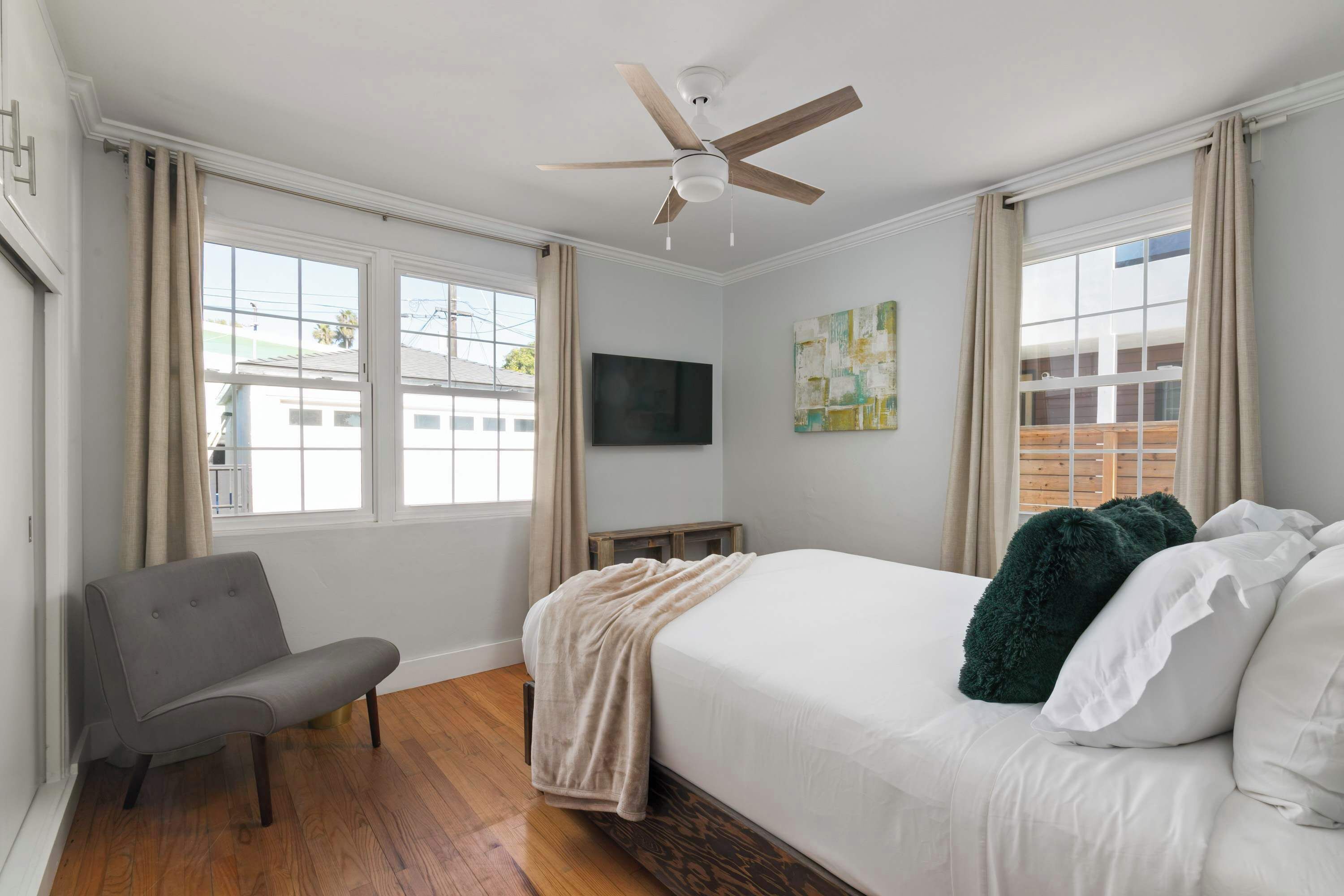 A bedroom with a bed, a chair, a ceiling fan, a TV, and a window with curtains.