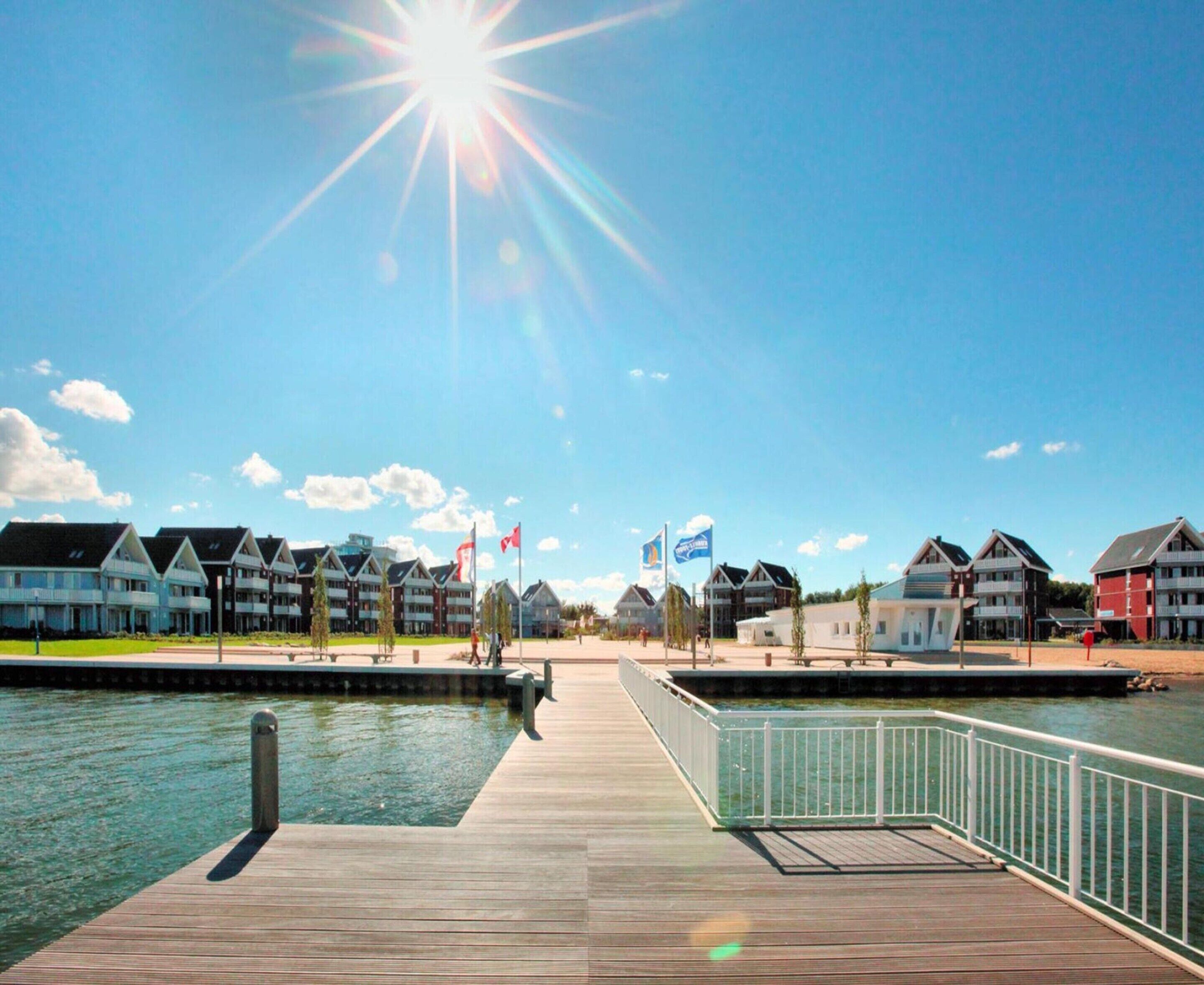 A wooden pier extending over calm water, leading to a row of houses with flags, under a clear blue sky.