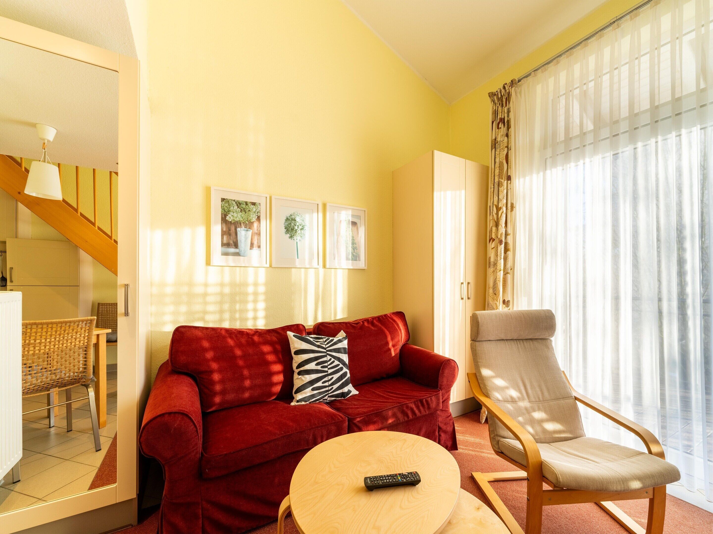 A living room with a red sofa, a wooden armchair, a round coffee table, and a remote control on it. There are three framed pictures on the wall, a staircase leading to the upper floor, and a large window with sheer curtains.