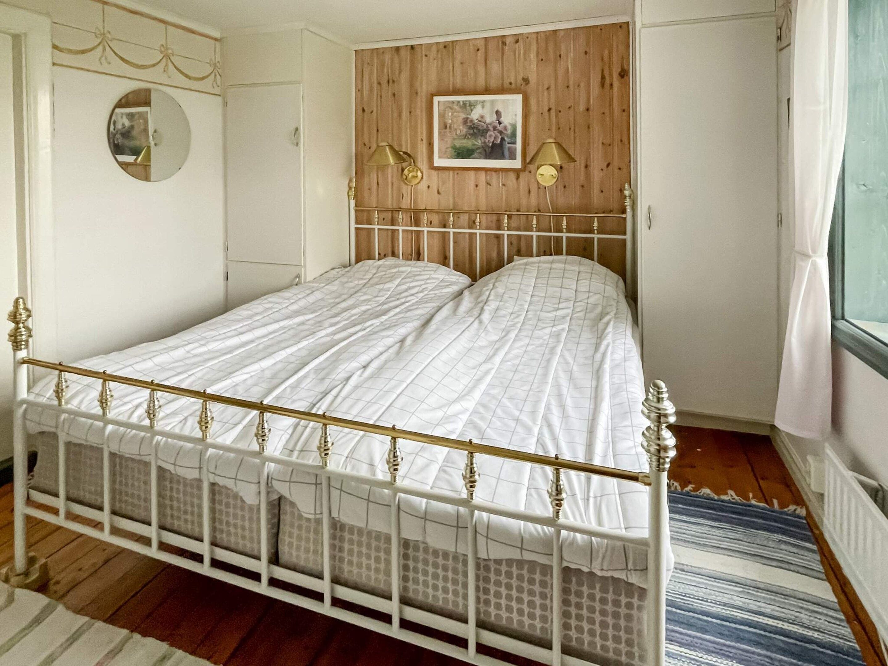 Une chambre avec un lit simple, une tête de lit en bois, un miroir rond, deux lampes fixées au mur et une fenêtre avec des rideaux.