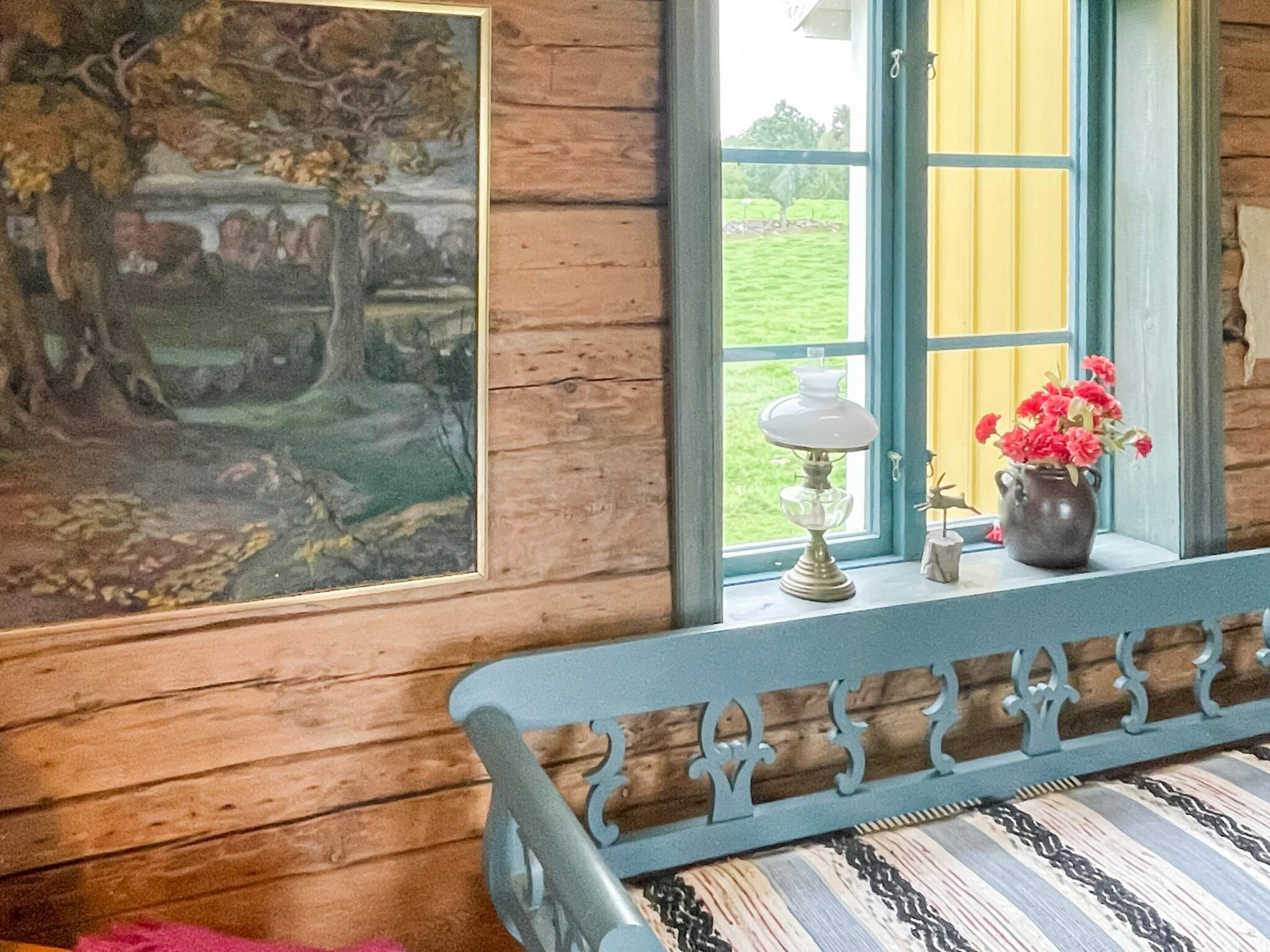 A room with a wooden bench, a painting, a window with a view of greenery, and a vase with flowers on the windowsill.