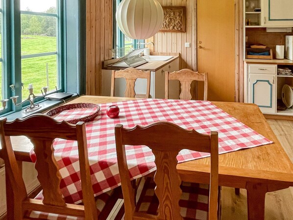 Un comedor con mantel a cuadros, sillas de madera, un ventanal con vistas a un paisaje verde, y una zona de cocina con armarios y un lavamanos.