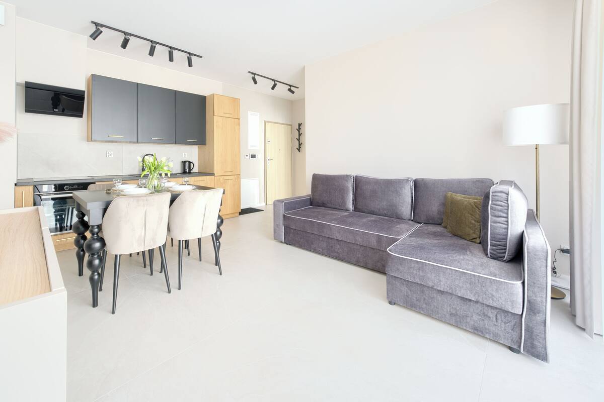 A modern living room with a grey sofa, a dining table with chairs, a kitchen area, and a flat-screen TV mounted on the wall.