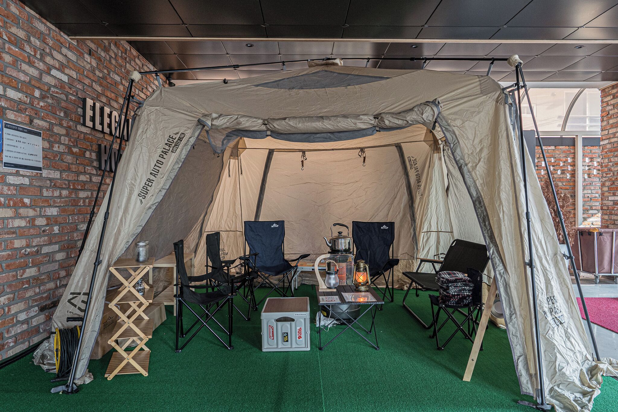 A tented area with a brick wall, a table with a kettle, chairs, and camping gear.