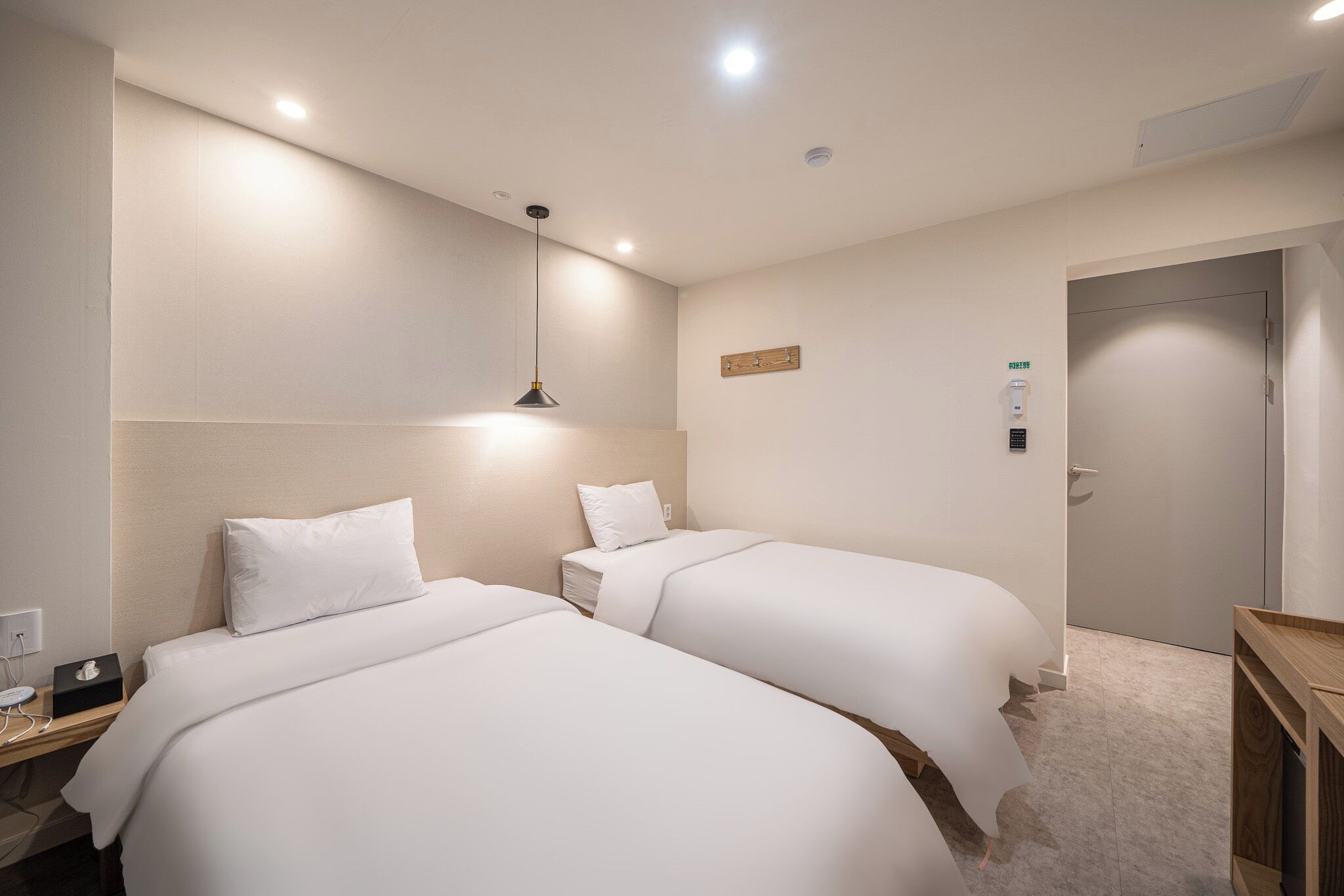 Une chambre d’hôtel avec deux lits, une table de chevet, une lampe, un panneau de commande fixé au mur et une porte.