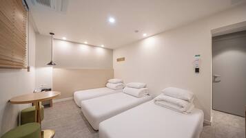 A hotel room with two single beds, a small table, a green chair, and a wall-mounted air conditioning unit.