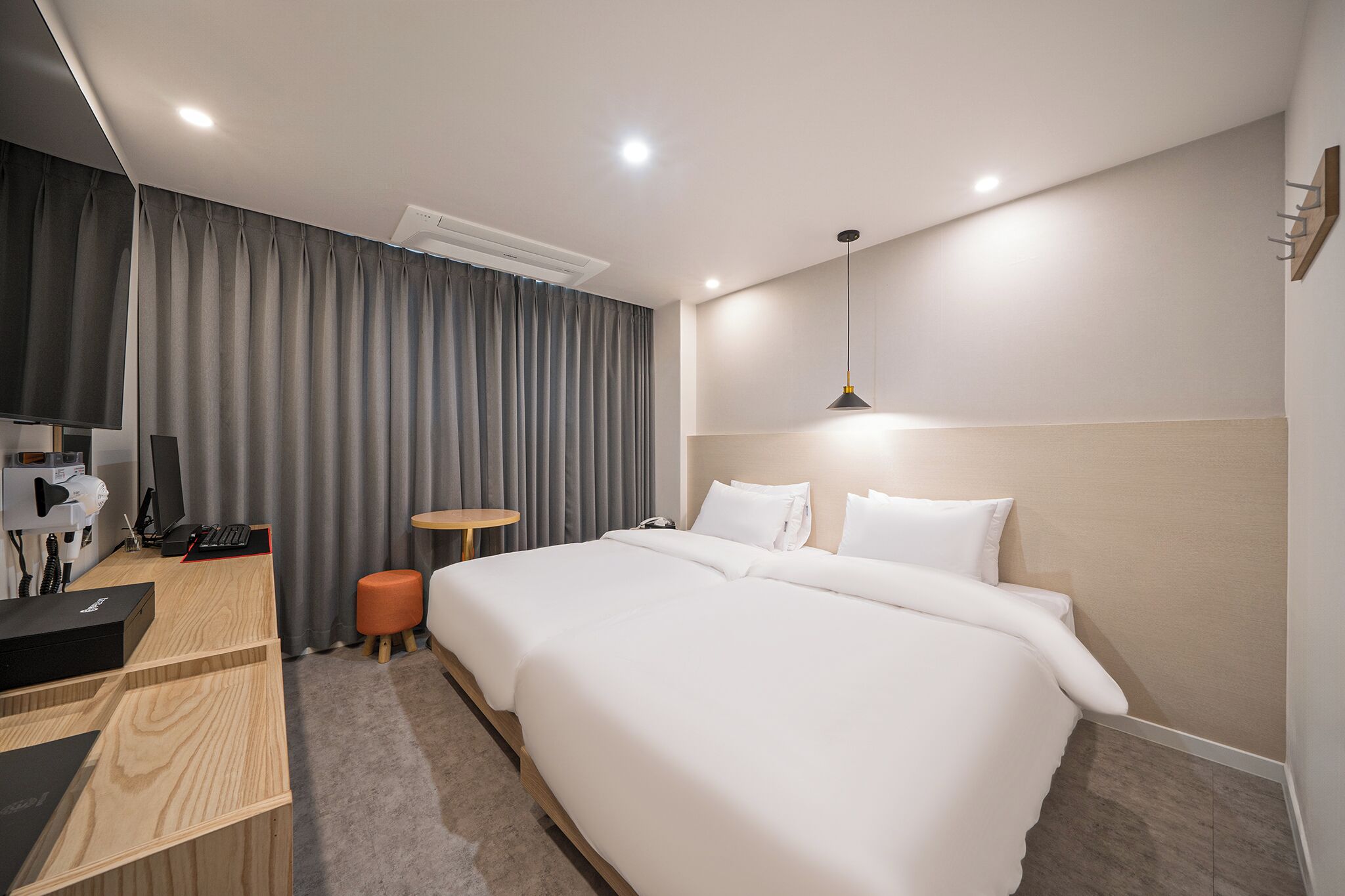 A modern hotel room with a large bed, a desk with a computer, a small stool, and a desk lamp.
