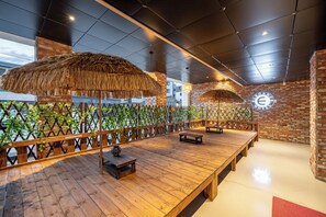 A spacious indoor area with a wooden deck, thatched umbrellas, and a brick wall with a logo. - Gyeongju Elegance Hwangridan-gil (Gyeongju)