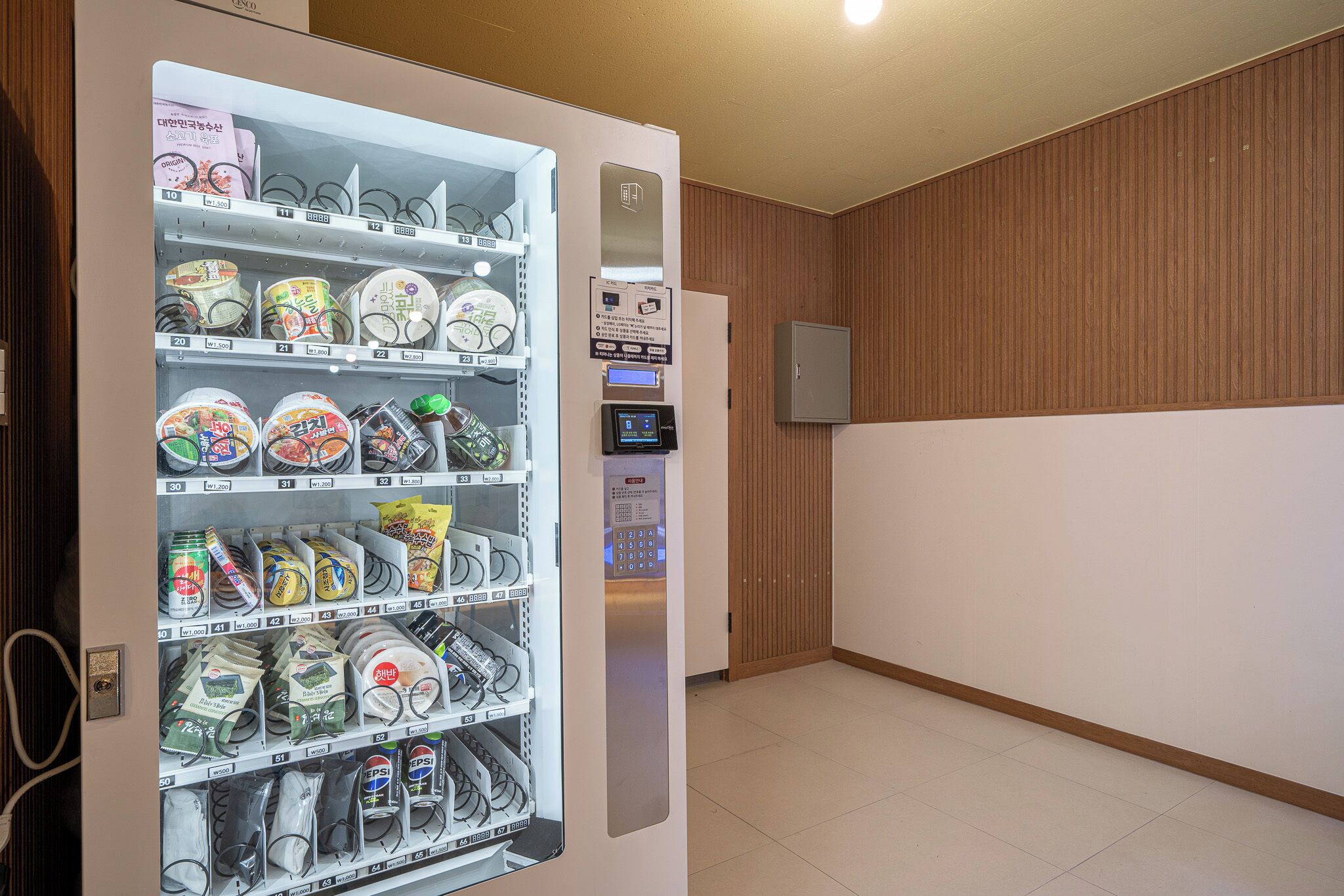 A vending machine with various packaged items, a digital payment kiosk, and a wooden panel wall.
