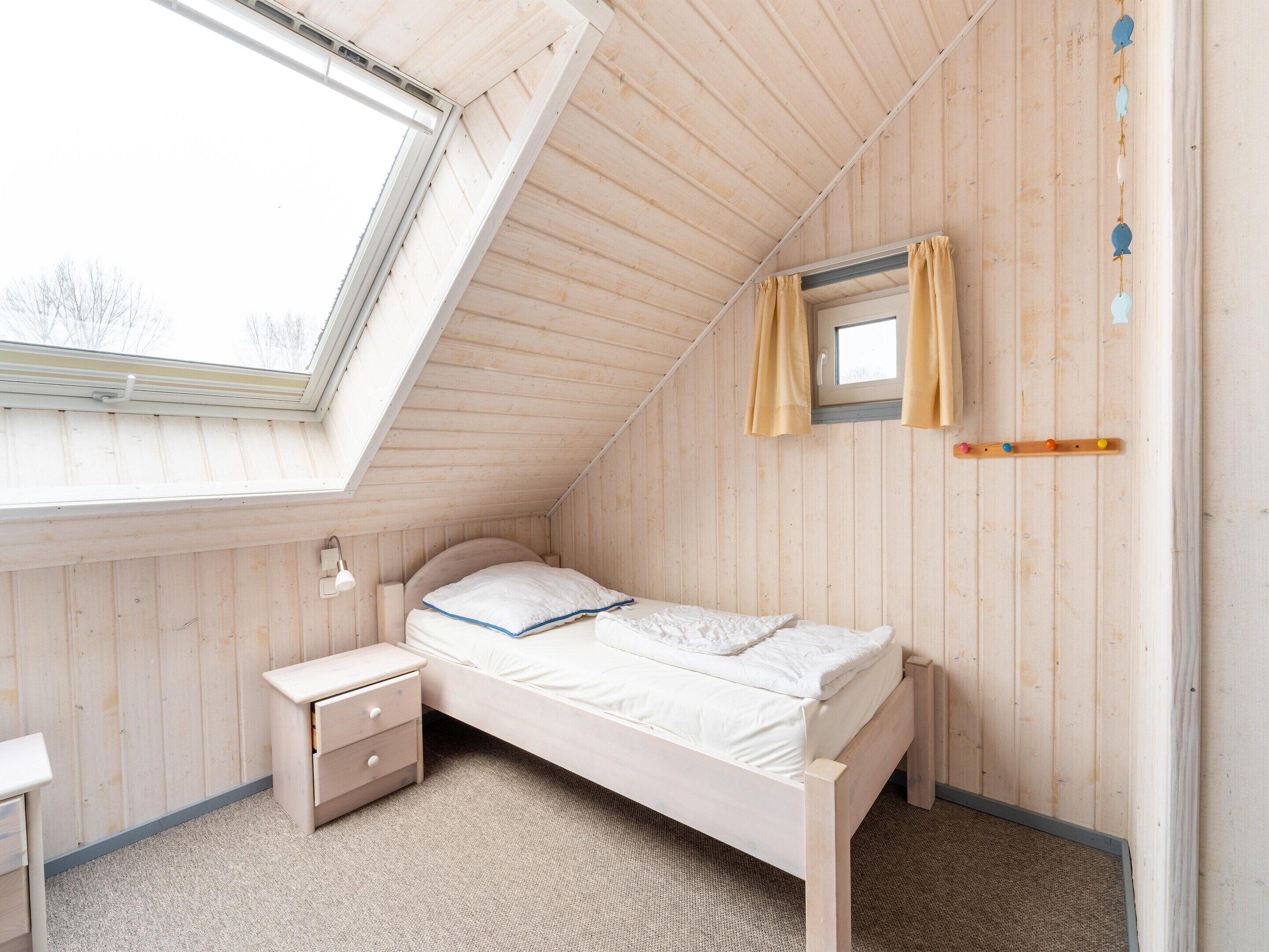 A small, cozy bedroom with a sloped ceiling, a single bed, a nightstand, a window with curtains, and a small wall-mounted shelf.
