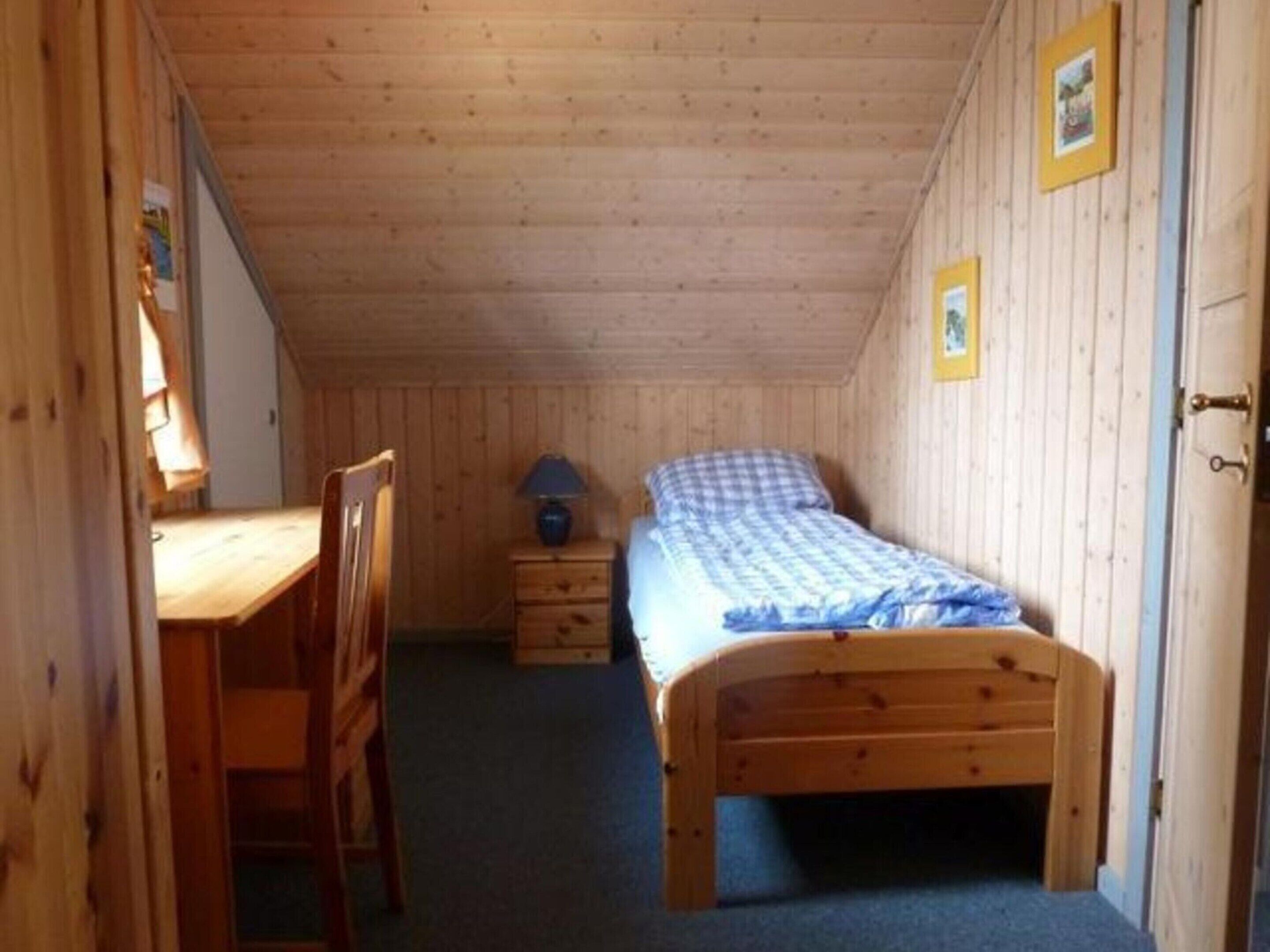 Ein kleiner Raum mit Holzwänden, einem Bett, einem Schreibtisch und einem Stuhl.