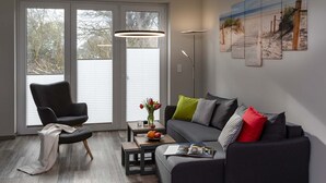 Ein modernes Wohnzimmer mit einem grauen Sofa, einem schwarzen Sessel und einem Couchtisch aus Holz. Große Fenster mit Jalousien, eine Stehlampe und ein Gemälde mit Strandmotiv schmücken den Raum.