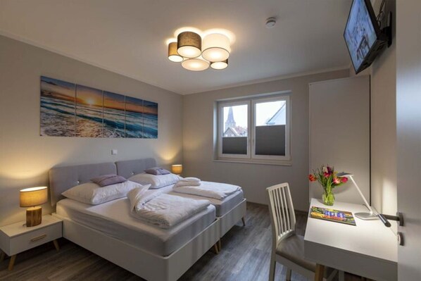 A bedroom with two beds, a desk with a chair, a TV, and a window with a view of buildings. - Exclusives Appartement auf Fehmarn (Fehmarn)