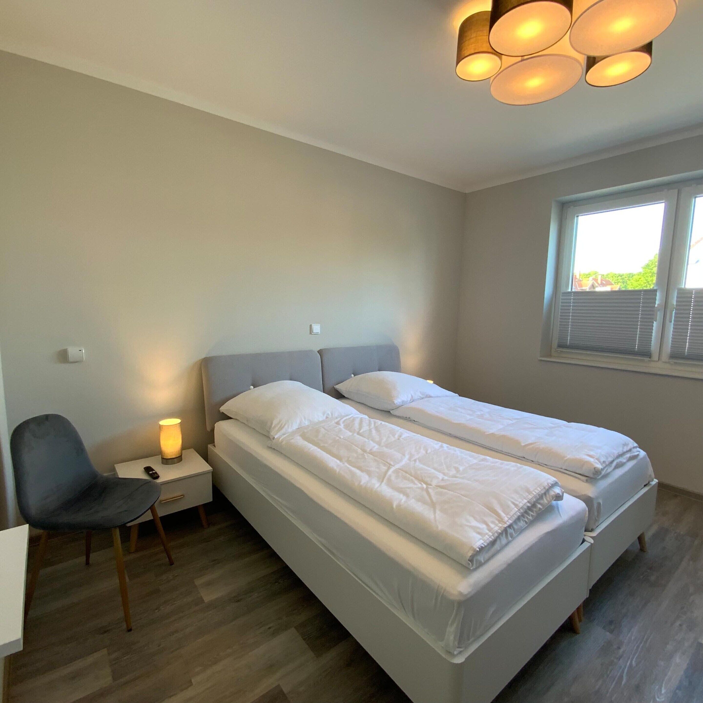 A bedroom with two beds, a bedside table with a lamp, a chair, and a window with blinds.