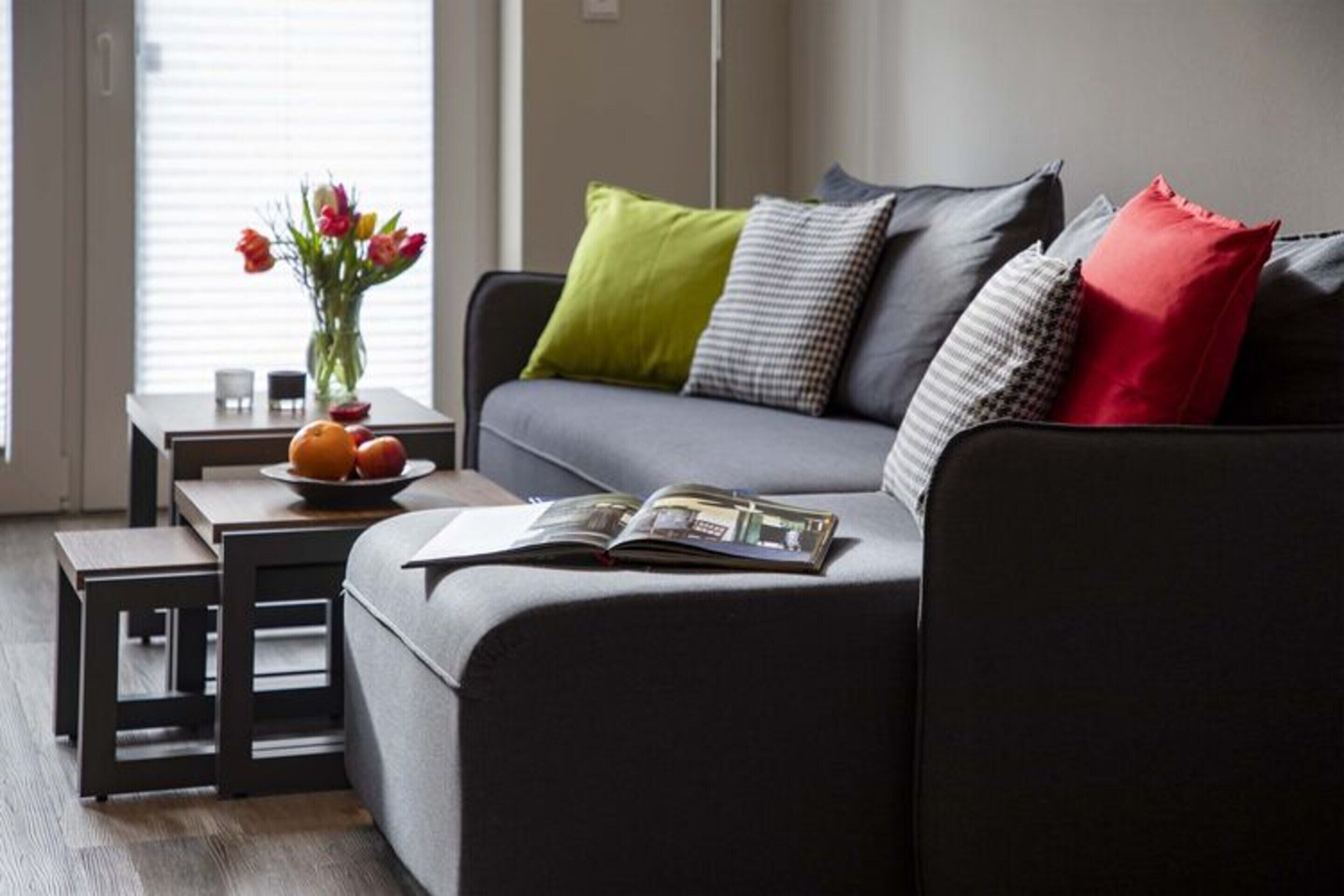 Ein modernes Wohnzimmer mit einem grauen Sofa, einem kleinen Tisch mit Blumen und einer Zeitschrift auf dem Sofa.
