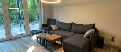 A modern living room with a grey sectional sofa, a wooden coffee table, and a black armchair. There is a glass door leading to a garden.