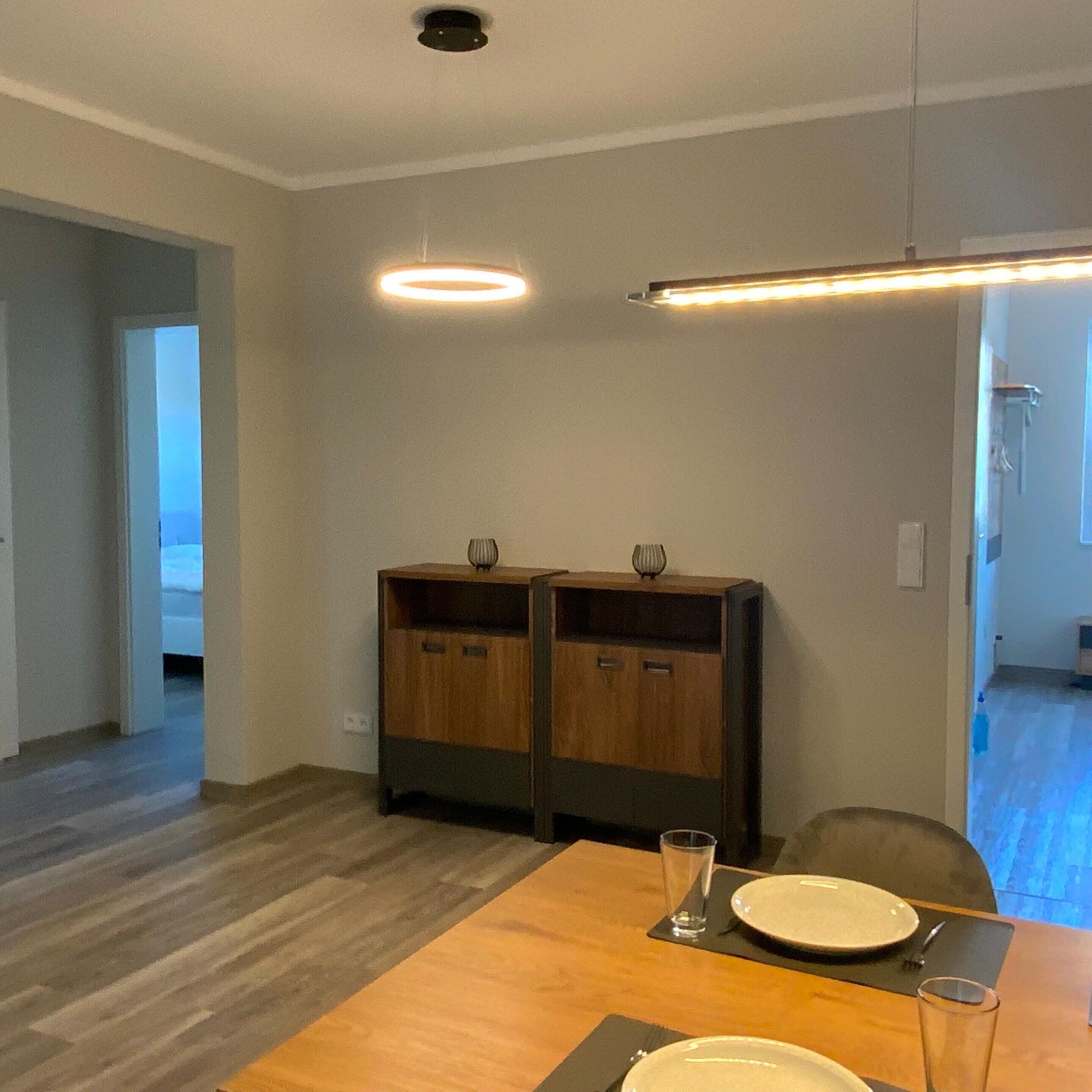 A dining area with a wooden table set for two, two wooden cabinets with handles, and a ceiling light fixture.