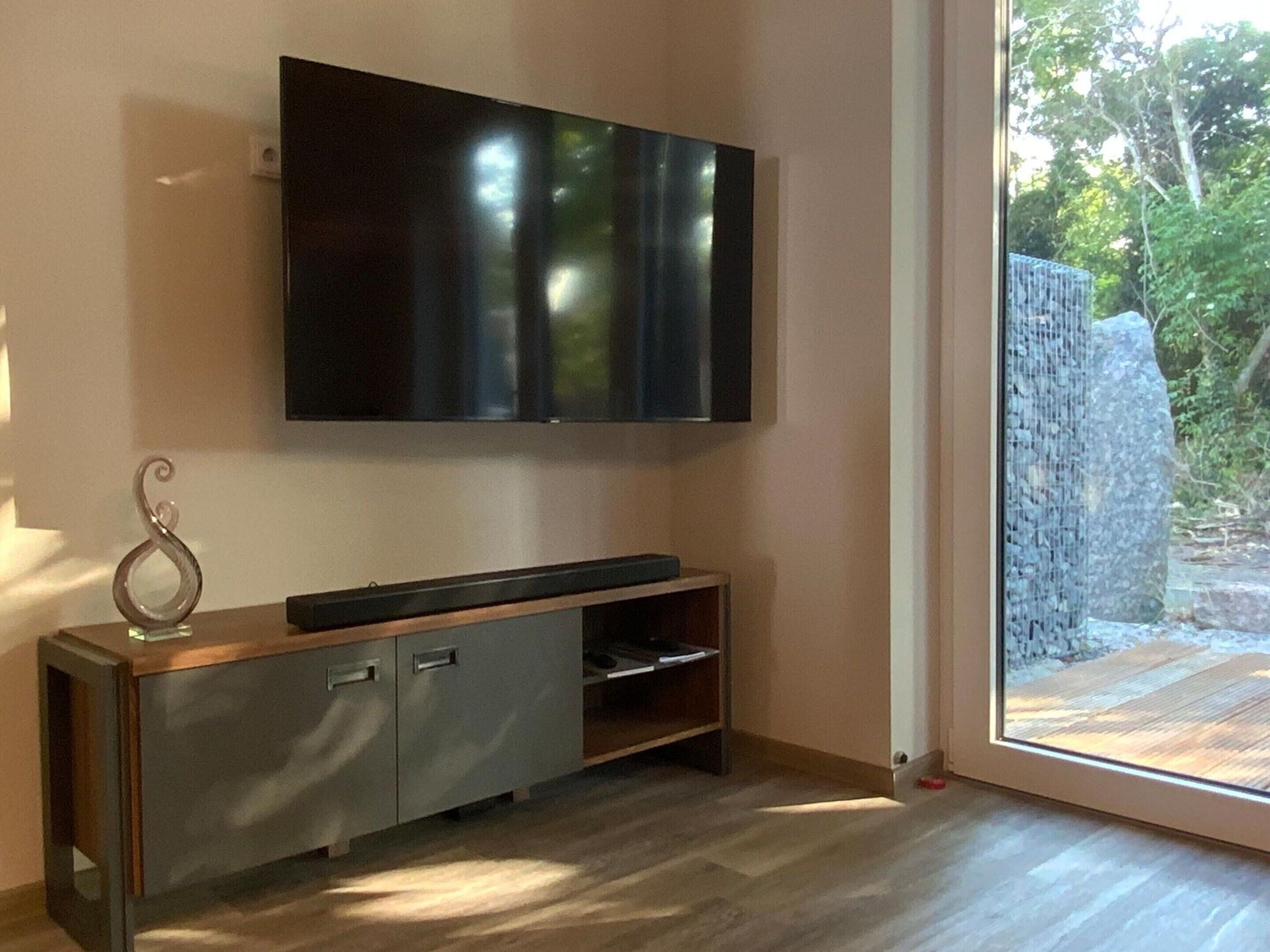 A modern living room with a flat-screen TV mounted on the wall, a wooden entertainment unit, and a large sliding glass door leading to an outdoor area.