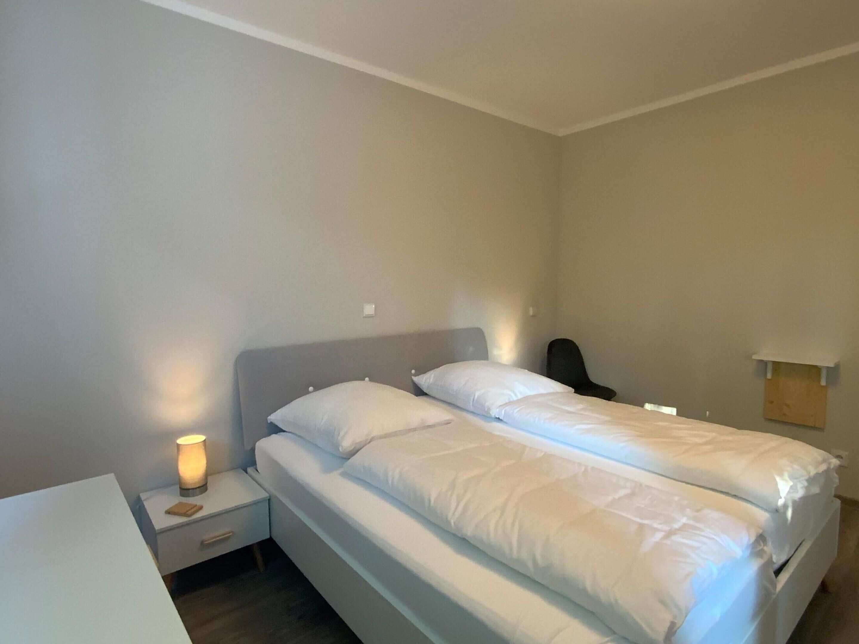 A single bed with white bedding, a bedside table with a lamp, and a chair in a simple room with plain walls.