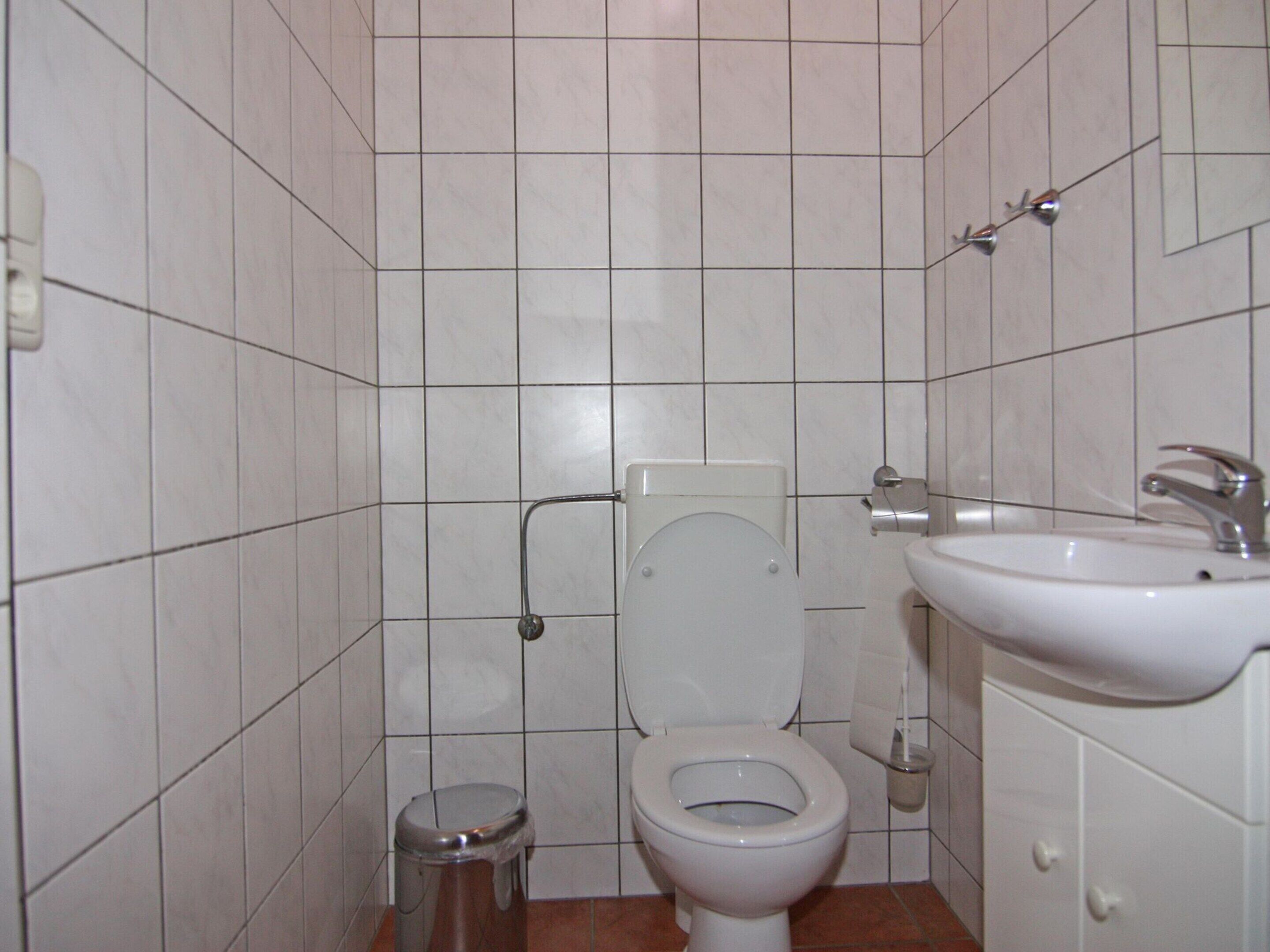 A small bathroom with white tiles, a toilet, a sink, and a trash can.