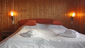 Una cama bien hecha con sábanas blancas y dos almohadas, frente a una pared de madera con dos lámparas de pared.