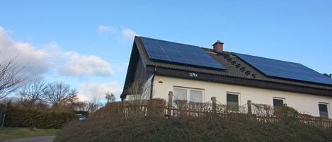 Een huis met een schuin dak bedekt met zonnepanelen, een witte gevel en een houten omheining.