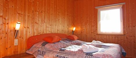 Una habitación de madera con una cama, dos lámparas fijadas a la pared, un ventanal y un banco de madera.