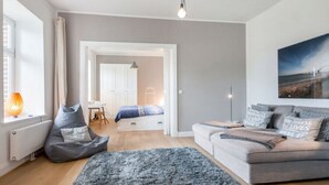 A modern living room with a sofa, a bean bag, and a rug. There is a bed with a blue blanket and a white headboard. A painting is on the wall, and a lamp is on the table. - Ferienhaus Kleine Freiheit auf Fehmarn (Fehmarn)