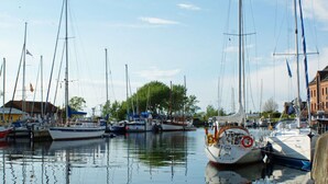 A marina with multiple sailboats docked, calm water, and a building with a red roof in the background. - Ferienhaus Kleine Freiheit auf Fehmarn (Fehmarn)