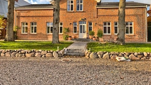 A two-story brick building with a symmetrical facade, a central entrance with a door and windows, and a gravel driveway with a stone border. - Ferienhaus Kleine Freiheit auf Fehmarn (Fehmarn)