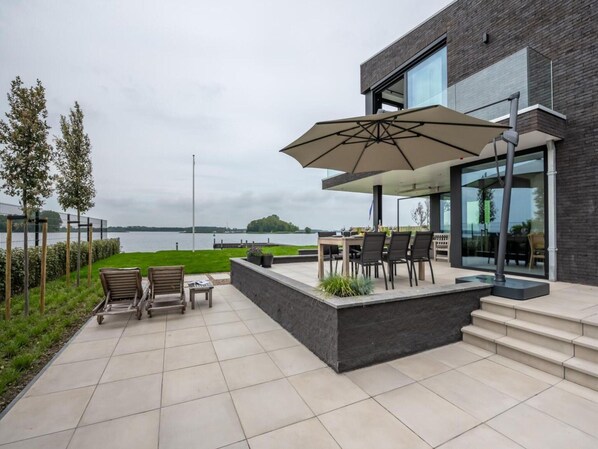 A modern outdoor patio with a view of a lake, a dining area with chairs and a table, and a large umbrella. - Attractive Villa With Jetty (Kamperland)