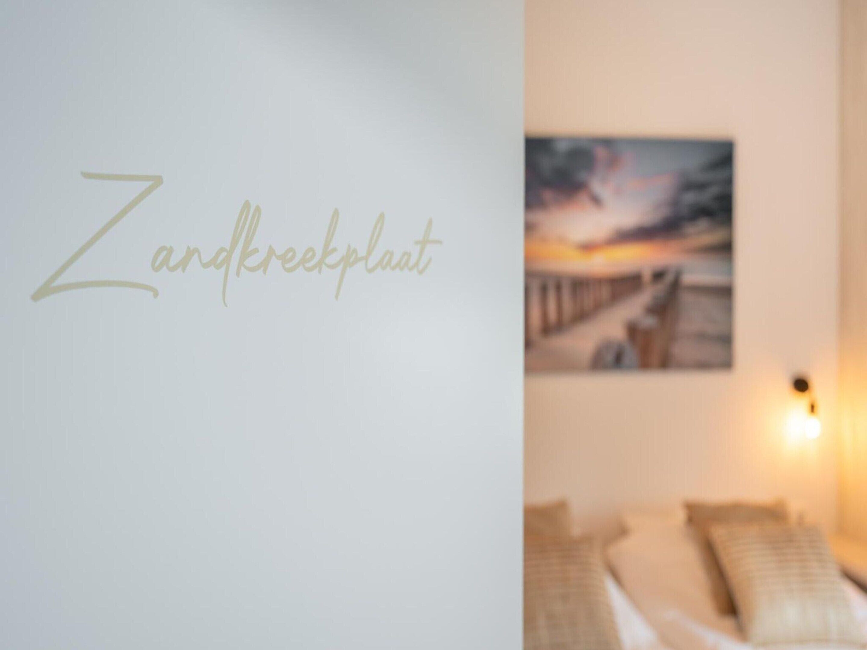 A room with a bed, pillows, a wall-mounted artwork, and a sign reading "Zandkrekplant".