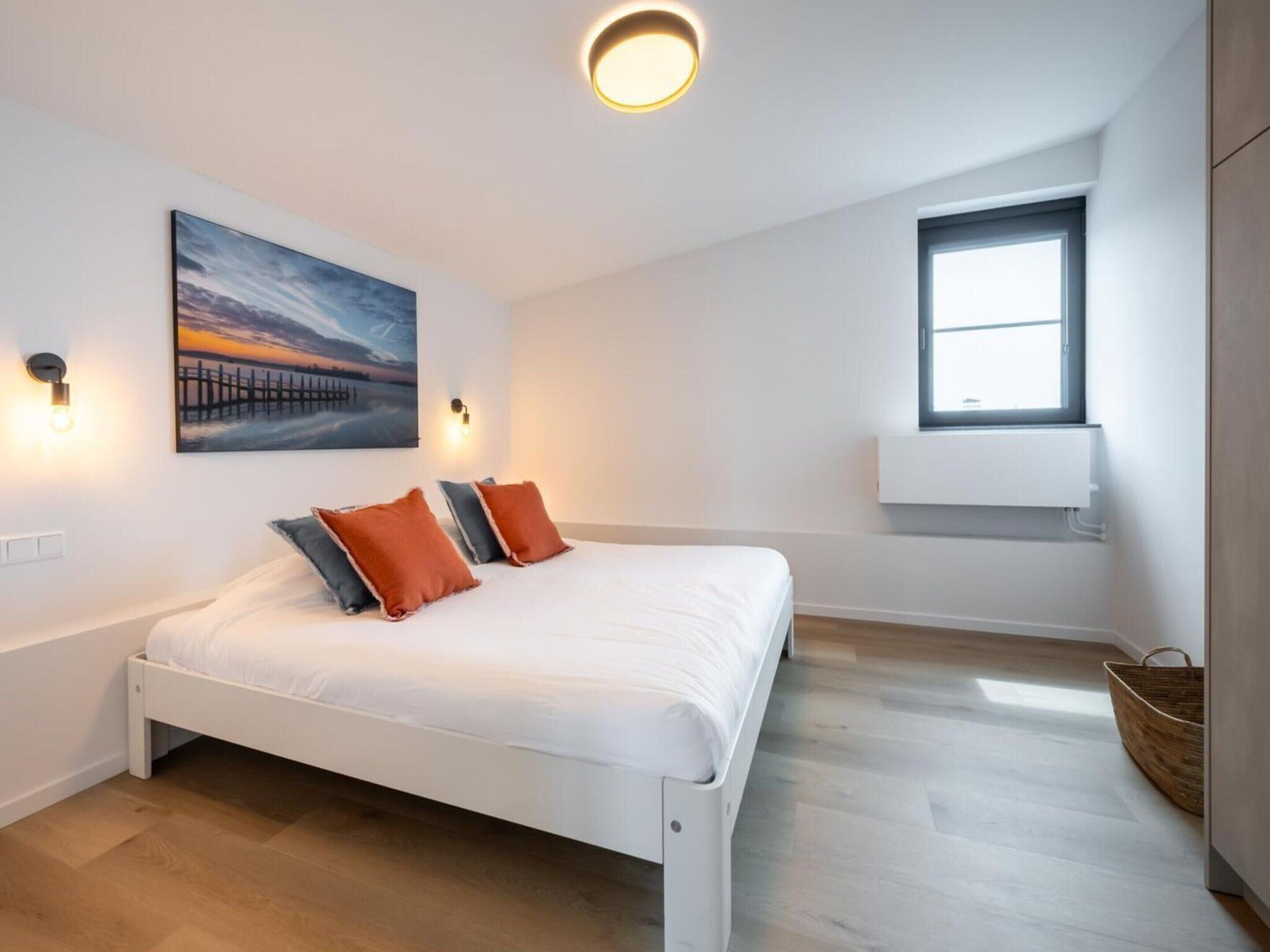 Une chambre à coucher moderne avec un lit blanc, deux oreillers, un tableau, une fenêtre et un luminaire au plafond.